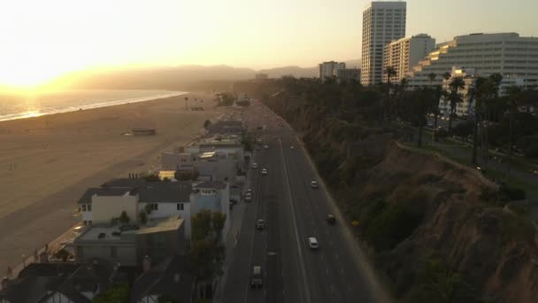 АЭРИАЛ: вид на скоростную автомагистраль Pacific Coast Highway HD рядом с Санта-Моникой Пьер, Лос-Анджелес с легким движением и видом на океан на закате волн, лето — стоковое видео