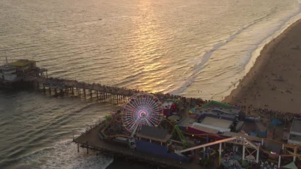 AERIAL: Κατεβαίνοντας στο Santa Monica Pier Ferrys Wheel, Λος Άντζελες στο όμορφο ηλιοβασίλεμα με τους τουρίστες, πεζοί με τα πόδια διασκεδάζοντας στο θεματικό πάρκο rollercoaster με κύματα θέα στον ωκεανό συντρίβεται — Αρχείο Βίντεο