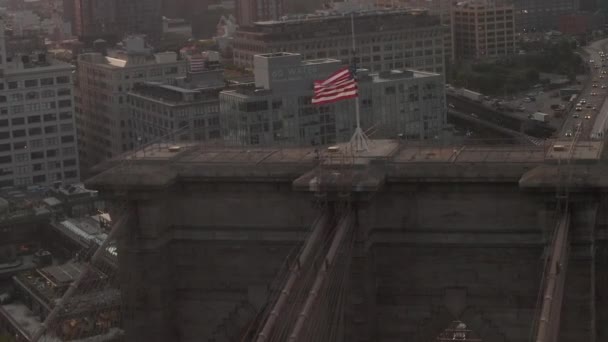 AERIAL:アメリカの国旗と霧のあるブルックリン橋の上のフライトを閉鎖するニューヨーク市スカイライン — ストック動画