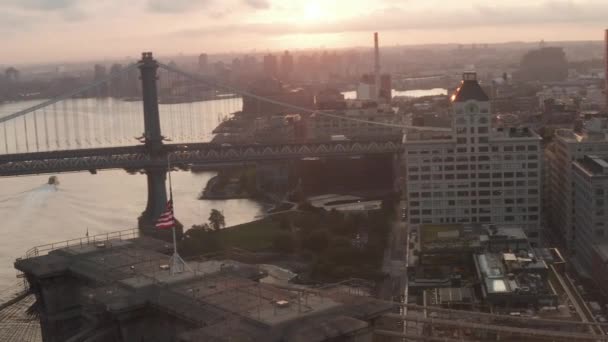 AERIAL: Närbild av Brooklyn Bridge med amerikansk flagga och vacker soluppgång morgon ljus över östra floden New York City — Stockvideo