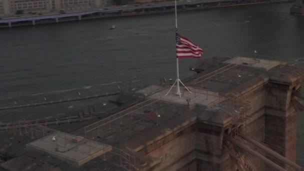 AERIAL: Närbild av Brooklyn Bridge med amerikansk flagga och vacker soluppgång morgon ljus över östra floden New York City — Stockvideo