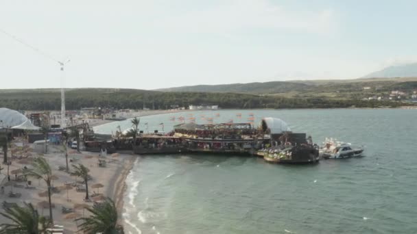 AERIAL: Piękny klub plażowy w Novalii, Chorwacja przy oceanie z falami w słoneczną pogodę i flagami, palmami i błękitną wodą — Wideo stockowe