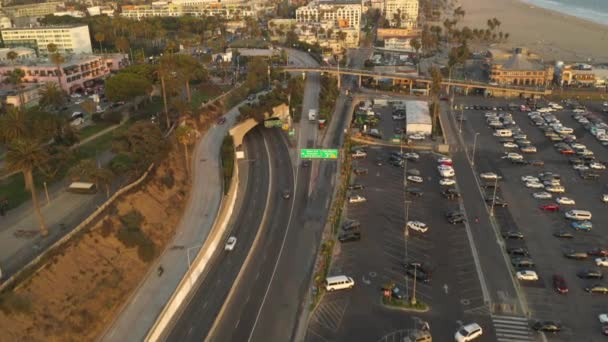 AERIAL: Voo sobre o Cais de Santa Mônica, estacionamento em Los Angeles, costa em direção ao cruzamento com o tráfego de luz e pássaros passando ao pôr do sol — Vídeo de Stock