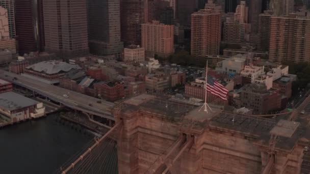 AERIAL: Close up voo círculo sobre Brooklyn Bridge com bandeira americana e East River vista Manhattan New York City Skyline — Vídeo de Stock