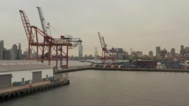Rıhtımda endüstriyel vinçlere doğru uçarken arka planda ve nehirde New York City silueti var. 