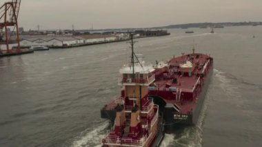 Bulutlu bir günde New York 'un rıhtımlarında büyük bir kırmızı kargo gemisini takip ediyoruz.