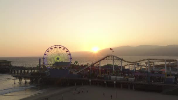 AERIAL: Vista de cerca de Santa Monica Pier Ferrys Wheel, Los Ángeles en el hermoso atardecer con turistas, peatones caminando divirtiéndose en la montaña rusa del parque temático con olas de vista al mar estrellándose — Vídeo de stock