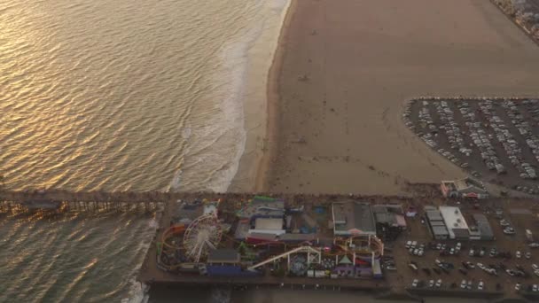 AERIAL: Flyger bort från Santa Monica Pier, Los Angeles vid vackra solnedgången med turister, fotgängare promenader har roligt på nöjespark berg-och dalbana med havsutsikt vågor kraschar — Stockvideo