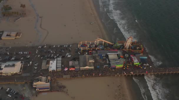 AERIAL: körözés Santa Monica móló, Los Angeles felülről gyönyörű naplemente a turisták, gyalogosok szórakozás vidámpark Ferrys Wheel óceánra néző hullámok összeomlik — Stock videók