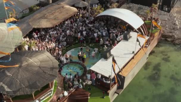 AERIAL: Close up of People dancing party at beachclub in Novalia, Croatia with ocean in sunny weather and flags, φοίνικες και πισίνες — Αρχείο Βίντεο