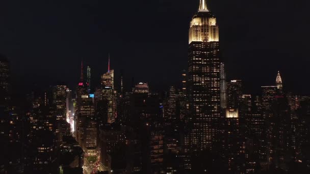 AERIAL: Vista de tirar o fôlego do icônico Empire State Building desaparecendo atrás de condomínios residenciais e edifícios de escritórios no centro de Manhattan, Nova York à noite — Vídeo de Stock