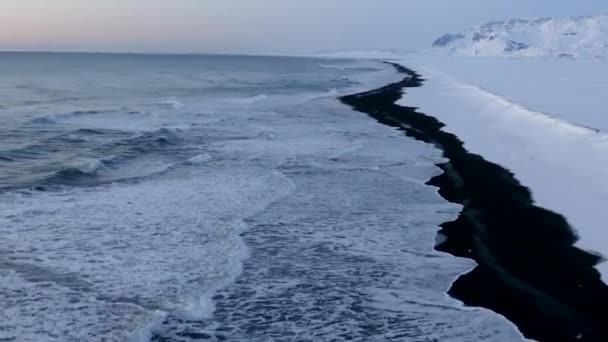 AERIAL: Voando sobre Black Beach com montanhas brancas de neve ártica no fundo na Islândia no inverno Neve, Gelo, Ondas, Água — Vídeo de Stock