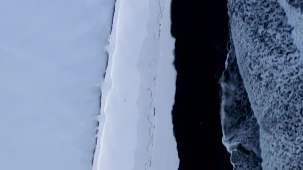 AERIAL:冬の雪、氷、波、水でアイスランドの風景、白い北極の雪の山と黒のビーチを見下ろす — ストック動画