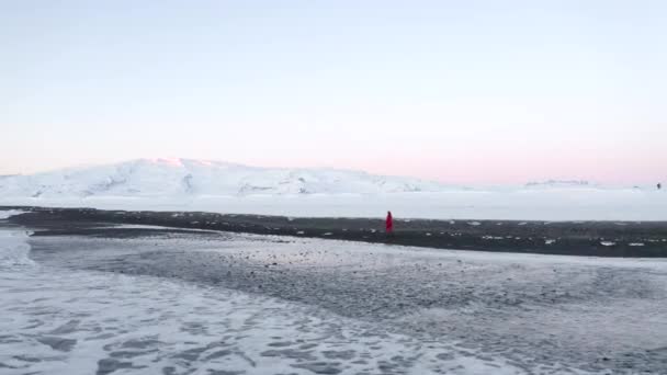 AERIAL: Drone tiro de pessoa andando em Black Beach com neve ártica branca na Islândia no inverno Neve, Gelo, Ondas, Água — Vídeo de Stock