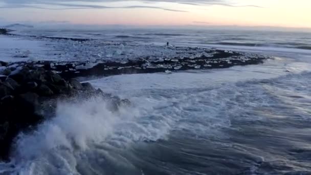 AERIAL:冬の雪、氷、波、水でアイスランドのブラック、ダイヤモンドビーチの岩に打ち寄せる波 — ストック動画