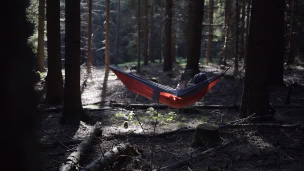 LOW MOTION: Jovem deitado em Orange Hammock entre duas árvores na floresta com bela luz de verão — Vídeo de Stock