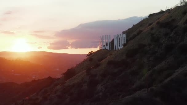 AERIAL:美しい夕日のハリウッドヒルズゴールデンアワーライトとカリフォルニア州ロサンゼルスのマウンテンサイドのハリウッドサインについての眺め — ストック動画