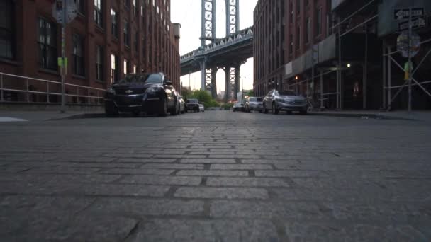 SLOW MOTION: Brooklyin Bridge Uitzicht vanaf Dumbo met dumbo appartementenhuizen en geparkeerde auto 's aan de zijkant in de zomer in de vroege ochtend licht — Stockvideo