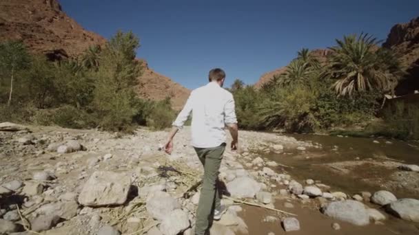 SLOW MOTION: YOUNG MAN WITH WHITE SHIRT WALKING OVER ROCKY BEACH NEXT TO RIVER ON BEAUTIFUL SUNNY DAY — Stock Video