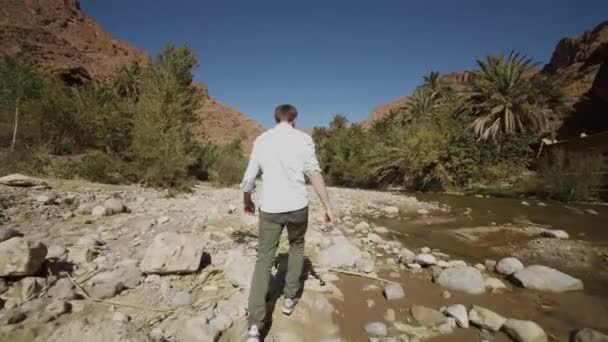 YOUNG MAN WITH WHITE SHIRT WALKING OVER ROCKY BEACH NEXT TO RIVER ON BEAUTIFUL SUNNY DAY — Stock Video