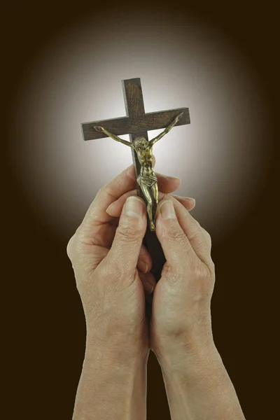 Praise the Lord - Female hands  holding a small statue of Christ on the Cross, against a dark brown background with white light behind the cross