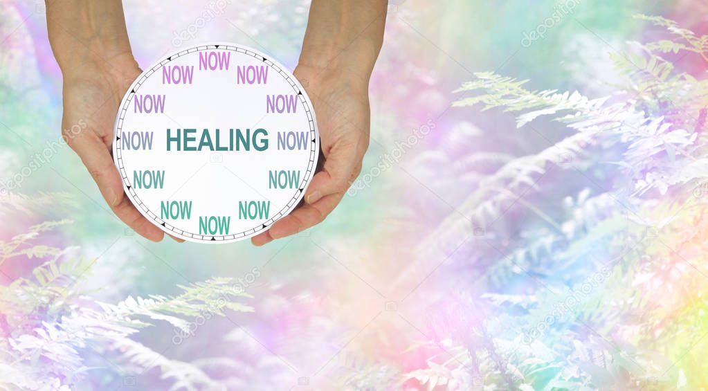 The time for healing is NOW - female hands holding a clock with no hands that has NOW in place of the numerals and HEALING instead of hands against a rainbow colored woods and fern background