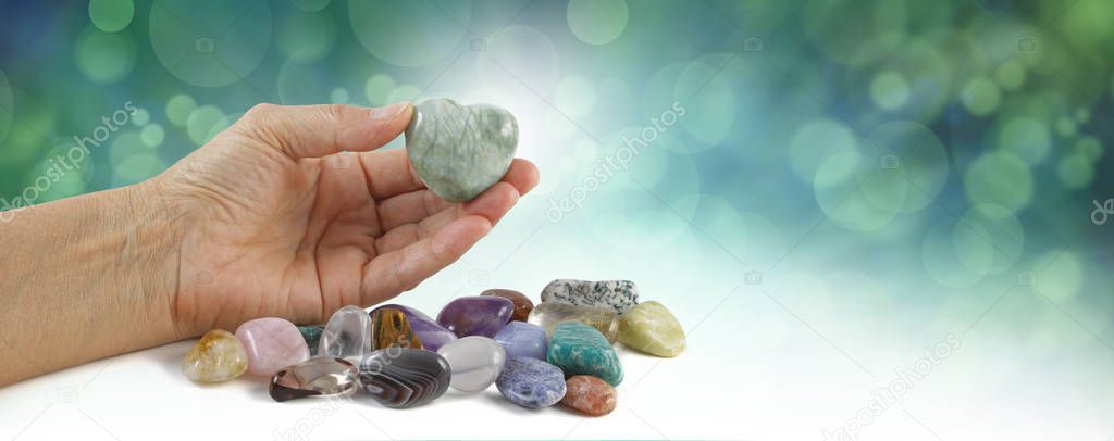 Crystal healer holding green puff heart stone - female hand offering a heart shaped crystal with a selection of other tumbled healing stones against a green bokeh background graduated to  white 