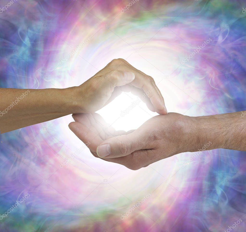 Our Combined energy is greater - female hand cupped over a male cupped hand with a bright white orb light between flowing out either side against a green and ruby red energy background with copy space