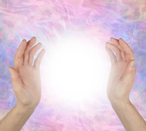 Sensing spiritual healing energy field between hands - female hands 25cm apart opposite each other with a bright white light orb energy between against a pale pink purple energy field  background