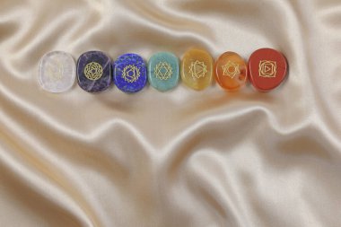 Single row of the Seven Major Chakras symbols - magenta, indigo, blue, green, yellow, orange and red polished flat gem stones etched with 7 chakra symbols on a gold silk material background with copy space clipart