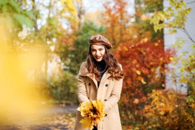 Şık bir kadın elinde sarı yapraklar tutuyor. Kadın bej ceketli, kahverengi şapkalı, bereli.. 