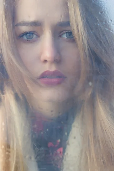 Beautiful sad girl with big eyes in a coat is behind wet glass — Stock Photo, Image