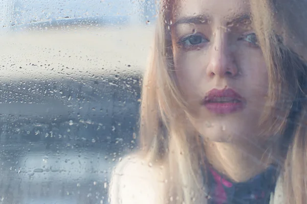 Beautiful sad girl with big eyes in a coat is behind wet glass — Stock Photo, Image