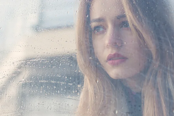 Menina triste bonita com grandes olhos em um casaco está atrás de vidro molhado — Fotografia de Stock