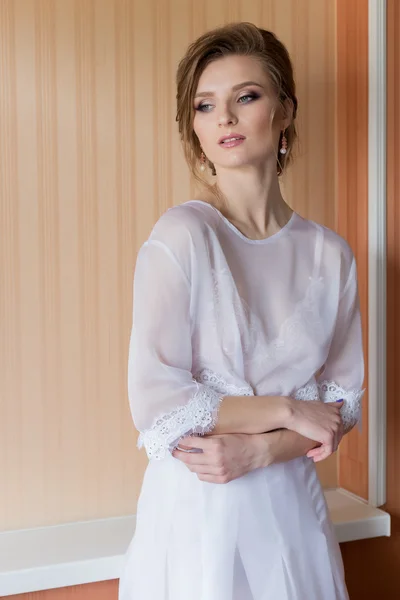 Manhã bela menina feliz da noiva perto de uma janela em um vestido branco com uma bela noite boudoir penteado festivo com ornamentos em seu cabelo e maquiagem macia — Fotografia de Stock