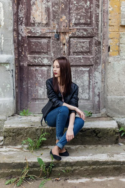 Красивая милая девушка моды с темными волосами в темных очках в кожаной черной куртке сидит на лестнице к крыльцу старых зданий в городе — стоковое фото