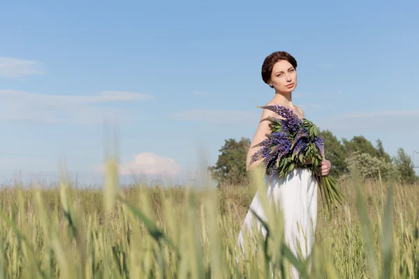 Hermosa chica dulce con un pelo trenzado en un vestido de verano blanco caminando en un campo con un ramo de flores de altramuz púrpura — Foto de Stock
