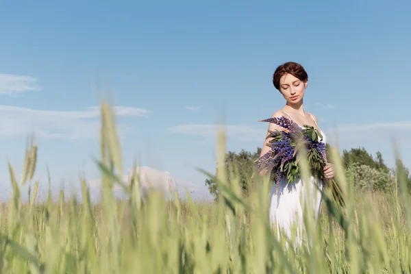 Красива мила дівчина з плетеним волоссям в білому літньому сараї, що йде в полі з букетом фіолетових люпинових квітів — стокове фото