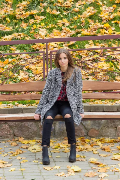 Bella ragazza dolce con i grandi occhi tristi in un cappotto seduto sulla panchina in autunno tra le foglie gialle cadute autunno giorno luminoso — Foto Stock