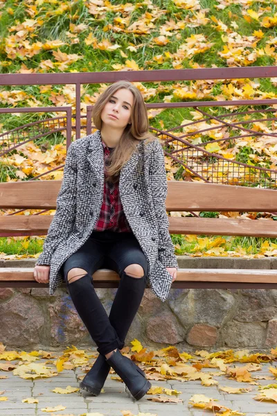 Красива мила дівчина з великими сумними очима в пальто, що сидить на лавці восени серед опалого жовтого листя осінній яскравий день — стокове фото
