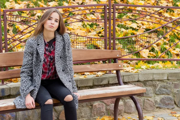 Piękna młoda dziewczyna z duże jesień smutne oczy w płaszcz i zgrywanie czarne dżinsy, siedząc na ławce w parku jesień — Zdjęcie stockowe