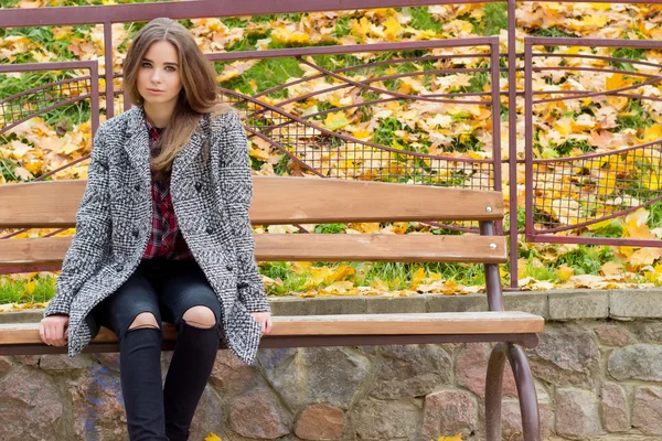 Mooi jong meisje met grote herfst droevige ogen in een jas en geripte zwarte jeans zittend op een bankje in de herfst park — Stockfoto