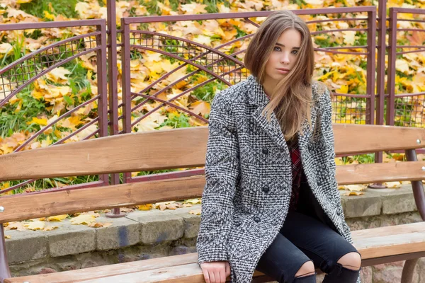 Красивая молодая девушка с большими осенними грустными глазами в пальто и разорванными черными джинсами, сидящая на скамейке в осеннем парке — стоковое фото