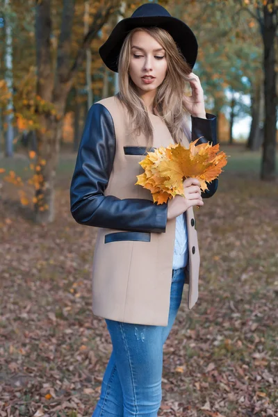 Красивая милая веселая девушка в черной шляпе с букетом ярких цветных листьев в пальто прогулки в осеннем парке — стоковое фото