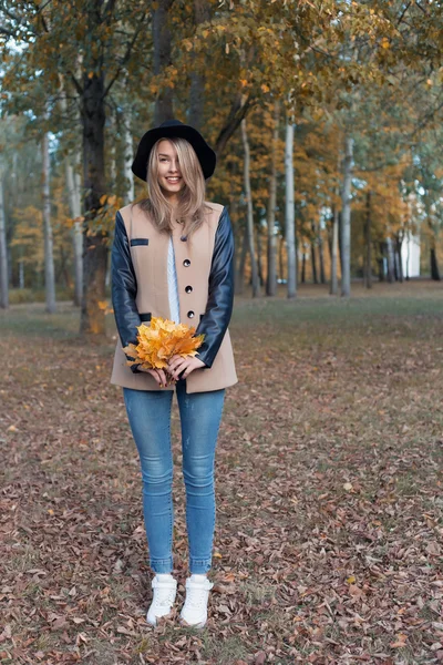 Красивая милая веселая девушка в черной шляпе с букетом ярких цветных листьев в пальто прогулки в осеннем парке — стоковое фото
