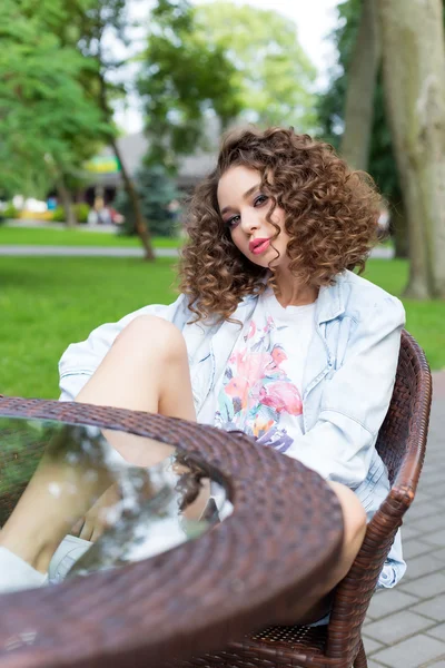 Beautiful cute cheerful girl in a bright dress with curly hair and bright make-up sitting at an outdoor cafe and waiting for coffee and ice cream — Stock Photo, Image