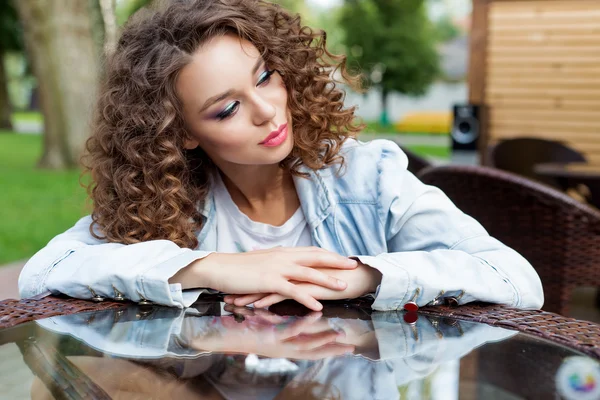 Hermosa joven con el pelo rizado se sienta en una mesa en un café al aire libre día de verano —  Fotos de Stock