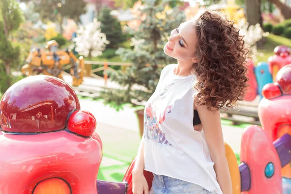 Beautiful young cheerful girl with curly hair in denim shorts and white T-shirt at an amusement park at sunset bright sun — Stock Photo, Image