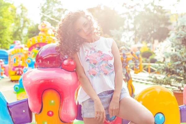 Beautiful young girl with curly hair in denim shorts and white T-shirt in the amusement park — Stock Photo, Image