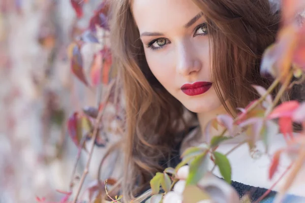 Autumn portrait of a beautiful woman with red hair with bright red lipstick on her lips in the city among the bright colored leaves on a sunny bright day — Stock Photo, Image